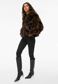 Brūna un melna mākslīgās kažokādas jaka ar teksturētu apdari un īsu dizainu, saskaņota ar melnajām šaurajām džinsām un melniem augstiem zābakiem.