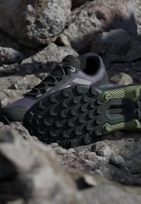 Svarta och lila sportskor med en robust svart sula och gröna detaljer, med textilmaterial, snören och förstärkt tåområde.