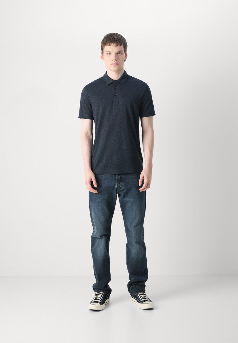 Junger Mann steht vor einer weißen Wand, trägt ein dunkelblaues Poloshirt, blaue Jeans und schwarz-weiße Turnschuhe, schaut nach vorne mit neutralem Ausdruck.