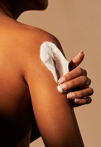 Crema idratante su una spalla liscia e abbronzata. La crema è spessa e bianca, in contrasto con il tono ricco della pelle.