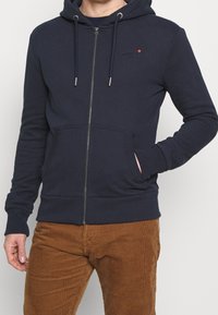 Hombre vestido con una sudadera con cremallera azul marino, con bolsillos frontales, y pantalones de pana marrón, con la mano en el bolsillo sobre un fondo claro y liso.