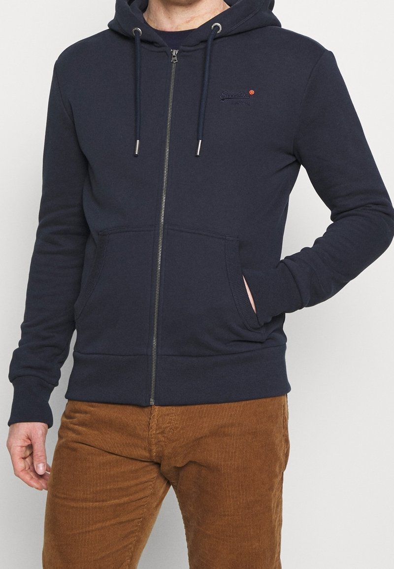 Hombre vestido con una sudadera con cremallera azul marino, con bolsillos frontales, y pantalones de pana marrón, con la mano en el bolsillo sobre un fondo claro y liso.