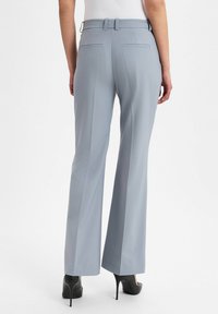 Frau in hellblauen maßgeschneiderten schmalen Hosen und schwarzen High Heels, die vor einem schlichten Hintergrund steht.