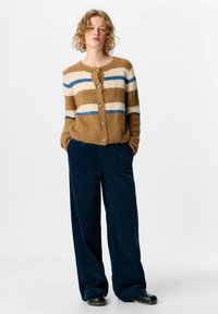 Frau mit lockigem blonden Haar, die eine braun-cremefarbene und blaue gestreifte Strickjacke, dunkelbraune weit geschnittene Cordhosen und schwarze, mit Nieten verzierte Schuhe trägt.