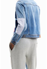 Person med en kortad blå denimjacka med randiga armbågsförstärkningar och ljusgråa sweatpants med fickor, händerna i fickorna.