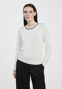 Biały dzianinowy sweter z czarnym ściągaczem przy dekolcie, długimi rękawami i dopasowanym krojem. Połączony z luźnymi czarnymi spodniami. Gładka tekstura.