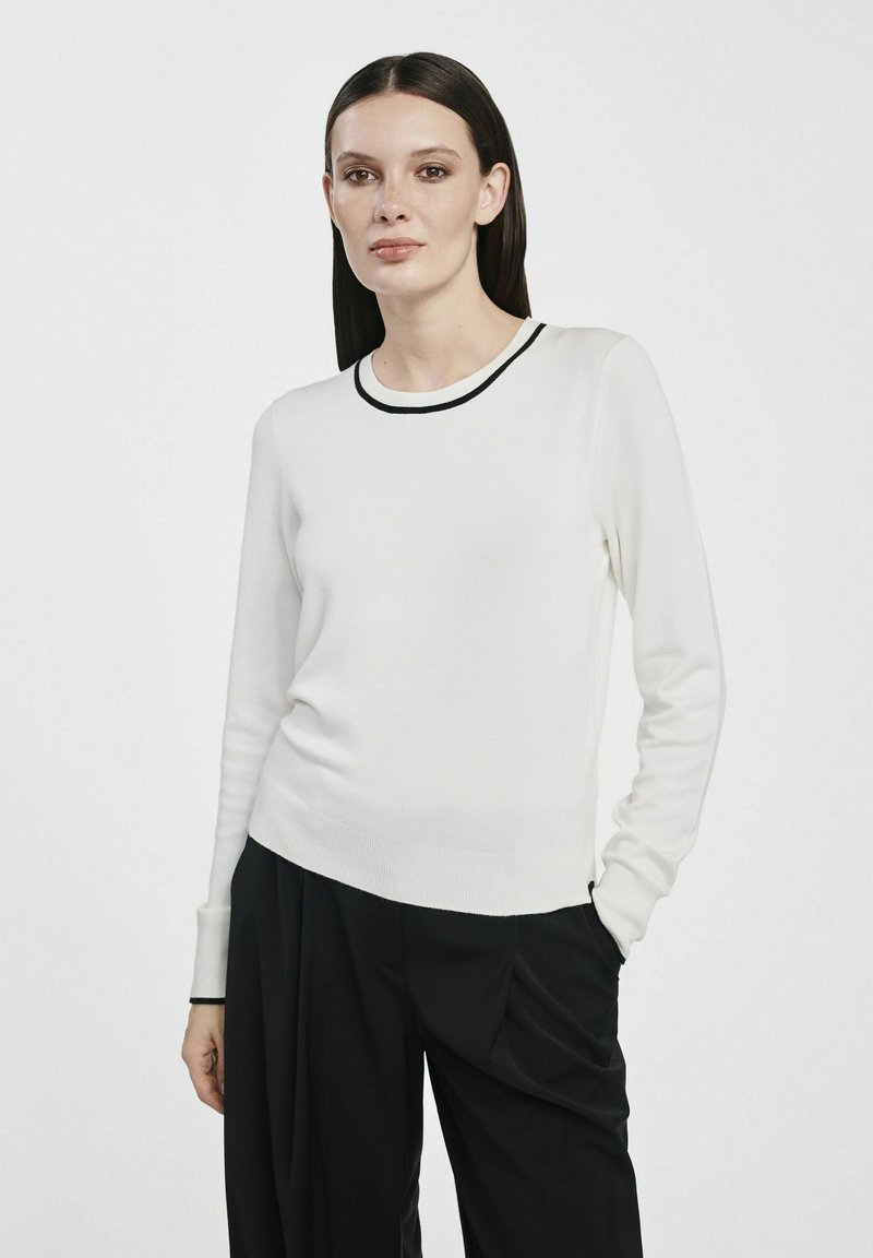 Biały dzianinowy sweter z czarnym ściągaczem przy dekolcie, długimi rękawami i dopasowanym krojem. Połączony z luźnymi czarnymi spodniami. Gładka tekstura.