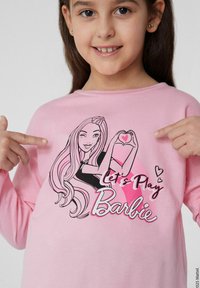 Světle růžová košile s dlouhým rukávem, na které je grafika kreslené holčičky s dlouhými vlasy, držící srdce. Text říká "Pojďme si hrát na Barbiu."
