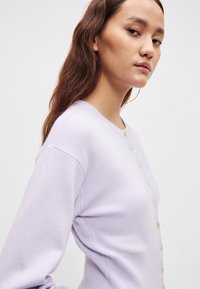 Λιλά ζακέτα κατασκευασμένη από απαλό πλεκτό ύφασμα, με ριμπ υφή, μακριά μανίκια και κλείσιμο με κουμπιά στο μπροστινό μέρος. Τα κουμπιά είναι ανοιχτού χρώματος.