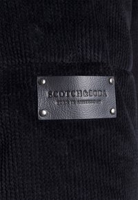 Tessuto nero testurizzato con un'etichetta in pelle che riporta "SCOTCH & SODA" e "NATO AD AMSTERDAM", fissata da borchie argentate.