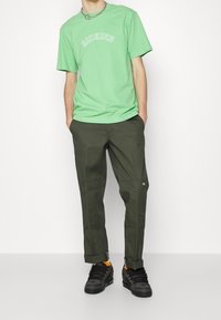 Grön bomullströja med "DICKIES"-text, ihopparad med mörk olivgröna byxor med en logotypapplikation. Svarta sneakers med orange detaljer fullbordar outfiten.