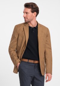 Ruskea bleiseri, joka on muotoiltu hyvin, päällä musta polo-paita. Harmaat housut ja ruskea rei'itetty vyö viimeistelevät asun.