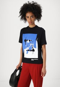 Crna majica kratkih rukava s plavim i bijelim grafičkim otiskom lica koje nosi sunčane naočale, s natpisom "KARL LAGERFELD JEANS."