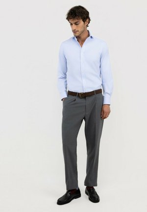 Hombre con camisa azul claro abotonada, pantalones grises con cinturón marrón y zapatos negros, de pie con las manos en los bolsillos frente a fondo blanco.