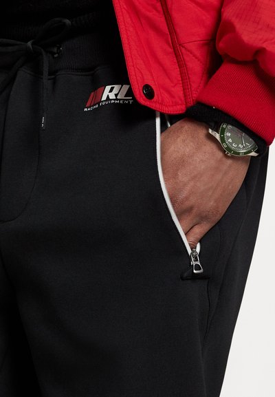 Pantalons de survêtement noirs avec un logo, des poches zippées et une taille élastique texturée, associés à une veste rouge et une montre verte.