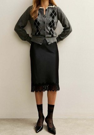 Femme portant un cardigan gris à motif losange, une jupe midi en satin noir avec un ourlet en dentelle, des chaussettes transparentes noires à pois et des talons pointus noirs.