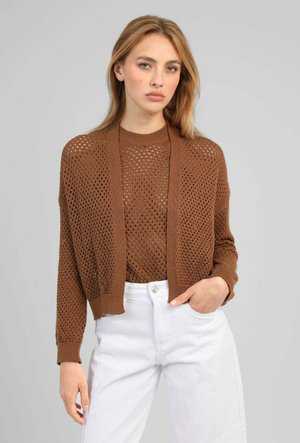 Jeune femme portant un cardigan marron en maille ouverte et un pull avec un pantalon blanc taille haute, debout avec une main dans la poche sur fond gris.