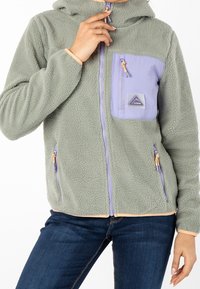 Chaqueta de forro polar en verde apagado con un bolsillo de parche en lavanda, cierre de cremallera y acentos en naranja. Tejido texturizado y diseño con capucha.