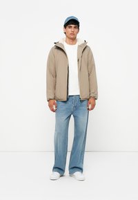 Beige pufferjakke med høj krave, lynlås-lukning og marineblå kasket, båret med en hvid t-shirt og lyseblå brede jeans. Hvide sneakers.