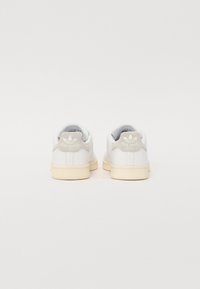 Zapatillas blancas de cuero con un soporte en el talón de un gris claro texturizado. Cuentan con una suela de goma crema y un logotipo sutil en el talón. Diseño clásico.