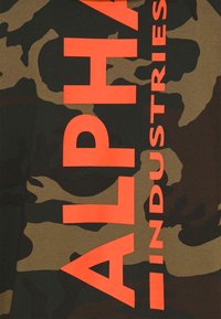 Zöld és barna árnyalatú terepszínű szöveten merészen, élénk narancssárga szöveg olvasható függőlegesen: "ALPHA INDUSTRIES".