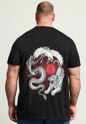 Mann trägt schwarzes T-Shirt mit weißem Drachen, Kranich und Tiger, die ein rotes Sonnenstrahl-Design auf dem Rücken umgeben, vor einfachem Hintergrund.
