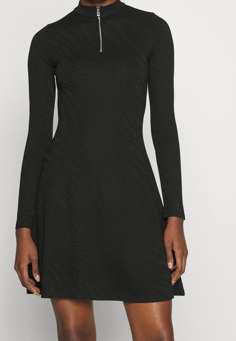 Robe noire à manches longues avec un col montant et une fermeture éclair à quart de zip. Présente un motif texturé sur toute la surface. Silhouette en A et longueur au-dessus du genou.