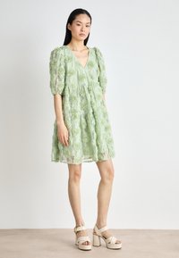 Robe en dentelle verte avec une texture florale, décolleté en V, manches bouffantes et jupe à niveaux, assortie à des talons plateformes beiges.
