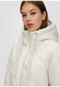 Kurtka puchowa w jasnym odcieniu kości słoniowej z pikowanym wzorem, wysokim kołnierzem i sznurkami do ściągania; posiada zamek błyskawiczny i akcenty z logo na wstążkach.