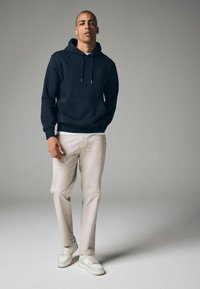 Sudadera azul marino con un bolsillo canguro, cordones y puños acanalados; combinada con pantalones beige claros y zapatillas blancas con detalles en gris.