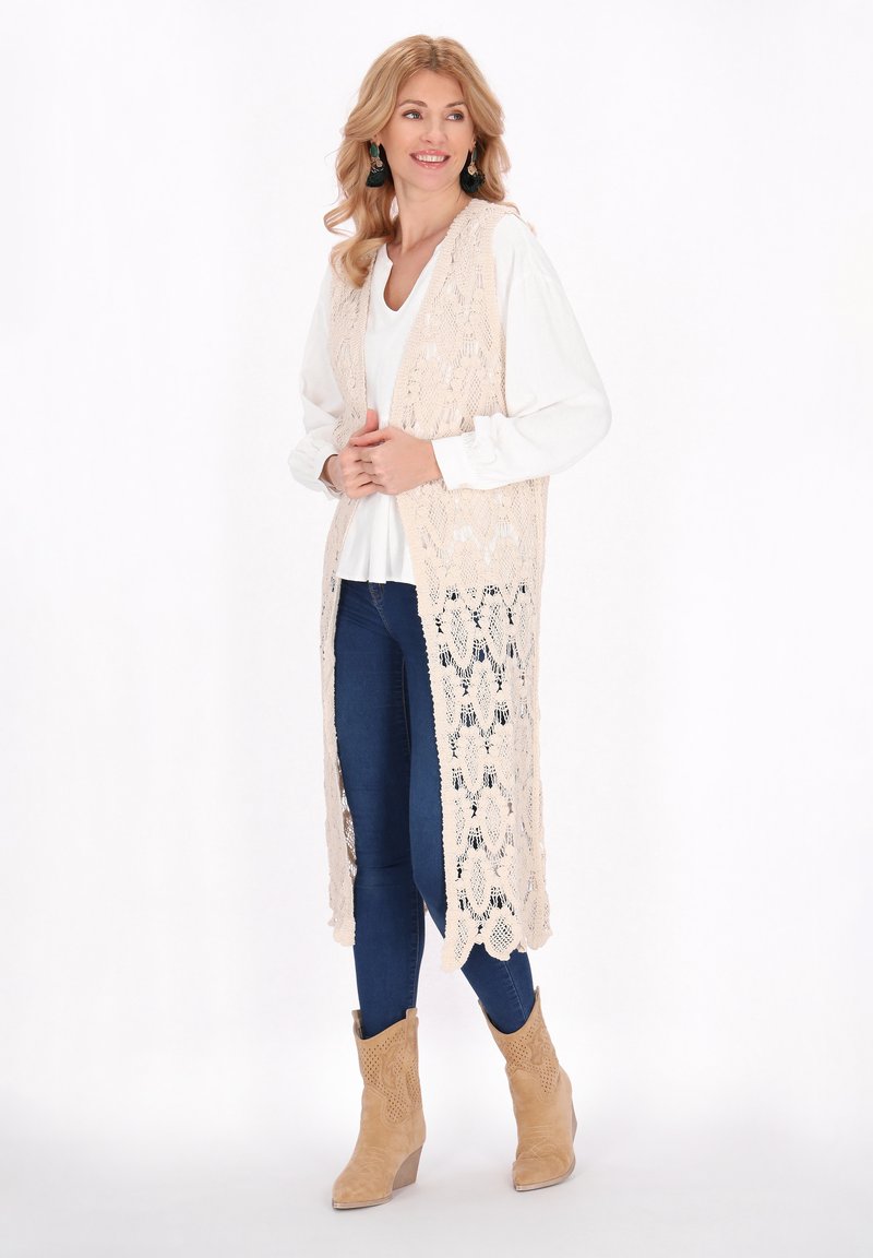 Beige Spitzenweste mit offenem Vorderteil, aufwendiges Muster, über einem weißen Top getragen, kombiniert mit blauen Jeans und beige Ankle Boots.