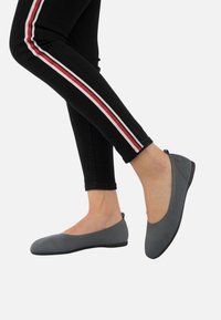 Chaussures plates grises avec une tige en tissu texturé, un bout arrondi et une semelle noire flexible, portées avec un jean noir présentant des rayures latérales blanches et rouges.