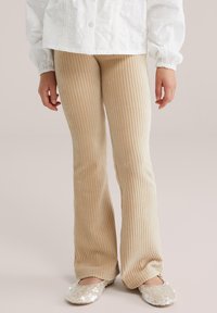 Bež hlače z rebrasto razširjenim krojem, izdelane iz mehke kordurove tkanine, v kombinaciji s svetlečimi svetlo obarvanimi čevlji brez vezalk. Model stoji z rokami sproščeno ob telesu.