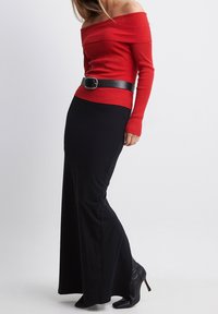 Pull rouge côtelé à manches longues et épaules dénudées, associé à une jupe longue noire et une large ceinture noire, complété par des talons hauts noirs.