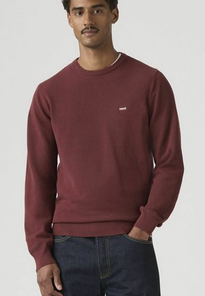 Mann trägt einen kastanienbraunen Levi's Rundhalspullover mit strukturierter Stoffstruktur und dunkelblaue Jeans, steht mit einer Hand in der Tasche vor einfarbigem Hintergrund.