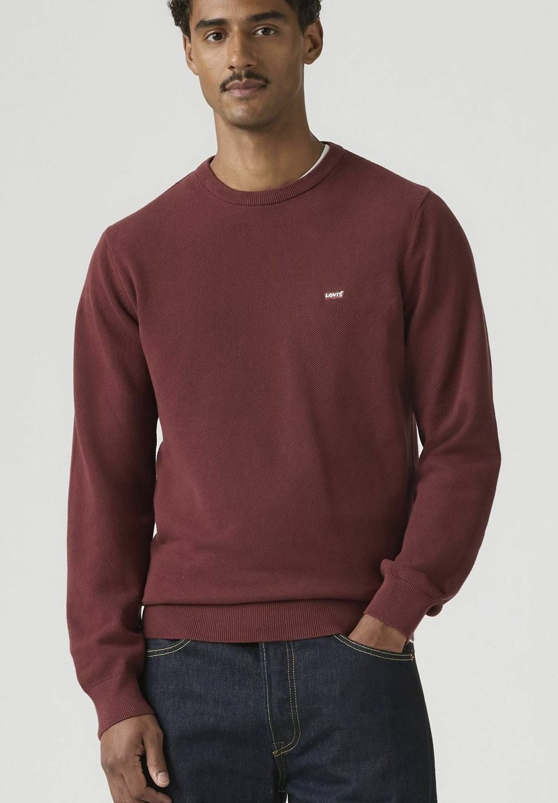 Hombre con suéter de cuello redondo color burdeos de Levi's con tela texturizada y jeans azul oscuro, de pie con una mano en el bolsillo contra un fondo liso.