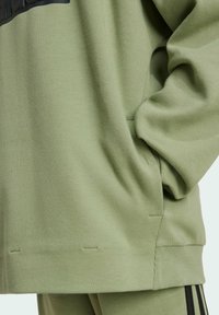 Felpa verde oliva con tessuto testurizzato, caratterizzata da una vestibilità ampia, tasca a marsupio e polsini a costine. Accenti neri sulla parte superiore.