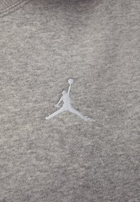 Grå hettegenser med en hevet, brodert hvit Jumpman-logo på forsiden, laget av et mykt, strukturert stoff.