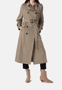 Mujer vestida con un abrigo trench de doble botonadura color beige con cinturón, pantalones negros de tiro corto y botines de ante color beige, de pie con las manos en los bolsillos.