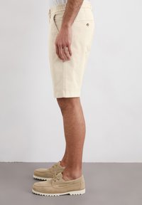 Beige cotton shorts with side pockets and an elastic waistband, featuring a back pocket with a metal button, displayed on a model's side.