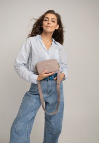 Borsa a tracolla in pelle beige con logo impresso, tracolla removibile e chiusura con zip, portata contro una camicia azzurro chiaro e jeans a stelle.