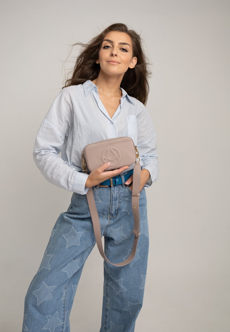Borsa a tracolla in pelle beige con logo impresso, tracolla removibile e chiusura con zip, portata contro una camicia azzurro chiaro e jeans a stelle.