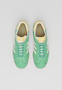 Sneakers in suede verde con strisce bianche, dettagli color crema e una soletta gialla con un motivo a logo ripetuto. Lacci piatti e suola in gomma.