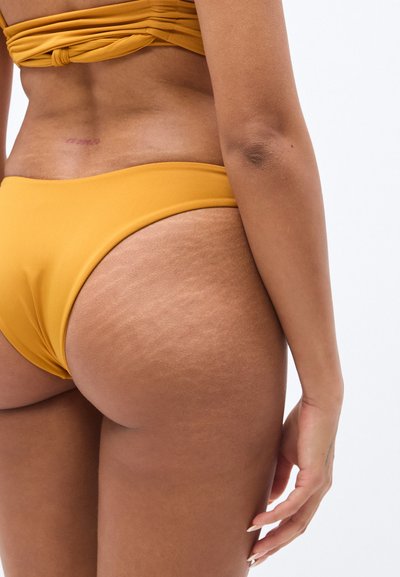 Κατώτερο μέρος μπικίνι σε μουσταρδί κίτρινο με ομαλή υφή, διαθέτει σχεδιασμό χαμηλής μέσης και ελάχιστη ζώνη στη  μέση, αποκαλύπτοντας το δέρμα και τις ραγάδες.