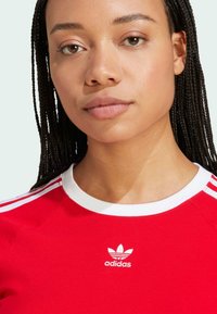 Red athletic shirt with white collar and three white stripes on shoulders. Features a small white Adidas logo on the chest.
