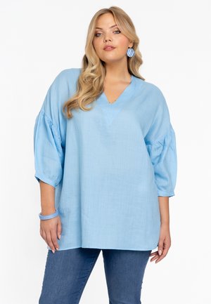 Himmelblaues, locker sitzendes Blusenshirt mit V-Ausschnitt und gep pufften Ärmeln, gefertigt aus einem weichen Material, kombiniert mit blauen Jeans.