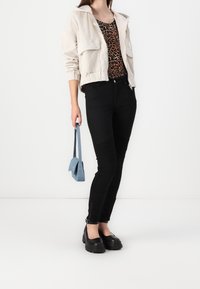 Kobieta w beżowej kurtce, topie w leoparda, czarnych spodniach i czarnych platformach, trzymająca małą niebieską torbę na tle białym.