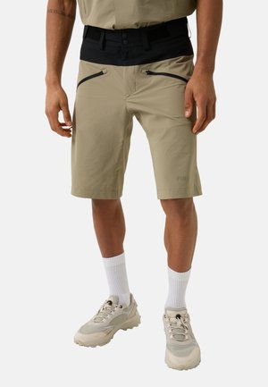 Beige Shorts mit schwarzem elastischem Bund, ausgestattet mit zwei Reißverschlusstaschen auf der Vorderseite und glatter Textur, kombiniert mit weißen Socken und hellen Sneakers.