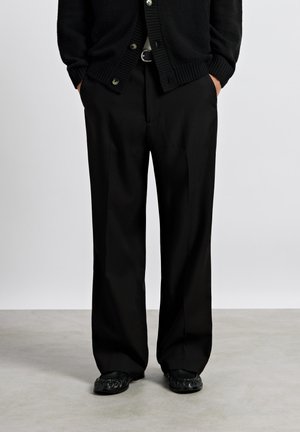 Personne portant un pantalon noir à jambes larges, un cardigan noir et des chaussures noires, debout les mains dans les poches sur un sol gris clair.