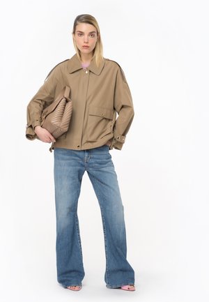 Giacca beige corta con bottoni dorati e due tasche frontali, abbinata a jeans in denim chiaro a palazzo, mentre tiene una borsa trapuntata color beige.